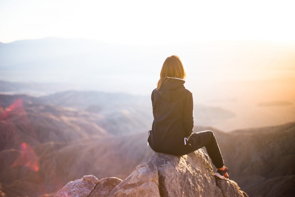 A woman sits on the precipice of a mountain, overlooking the valley below as the sun sets.