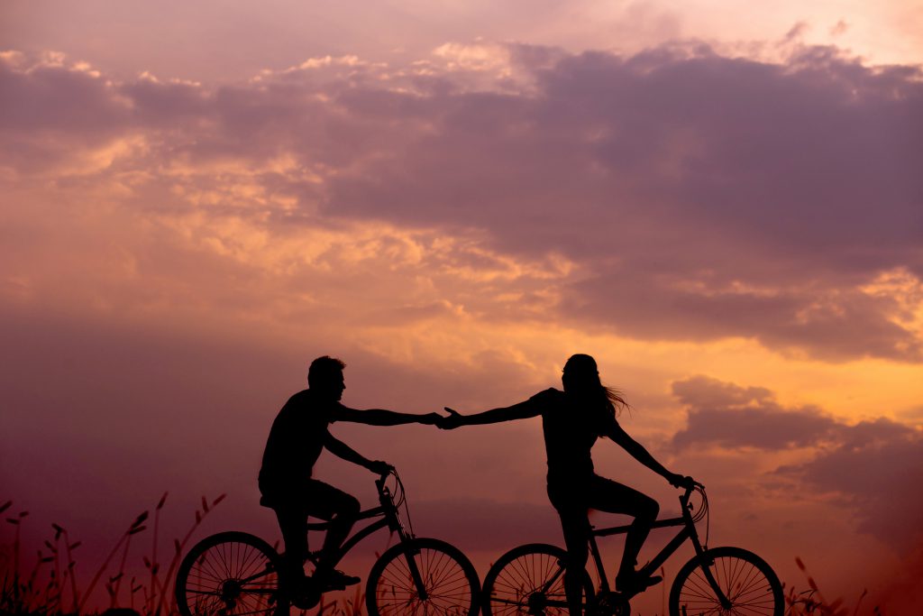 The silhouette of two people riding bikes with hands outstretched towards each other and a twilight sunset in the background.