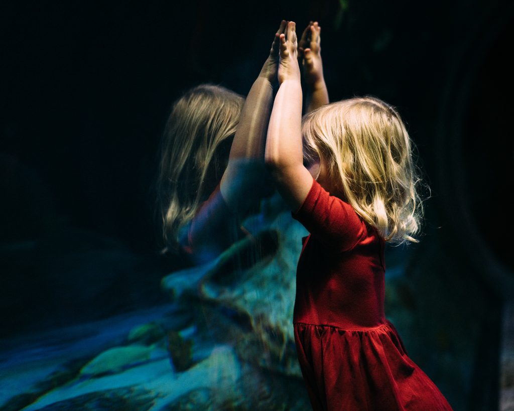 A little girl stands at the glass of an aquarium with her arms raised overhead.