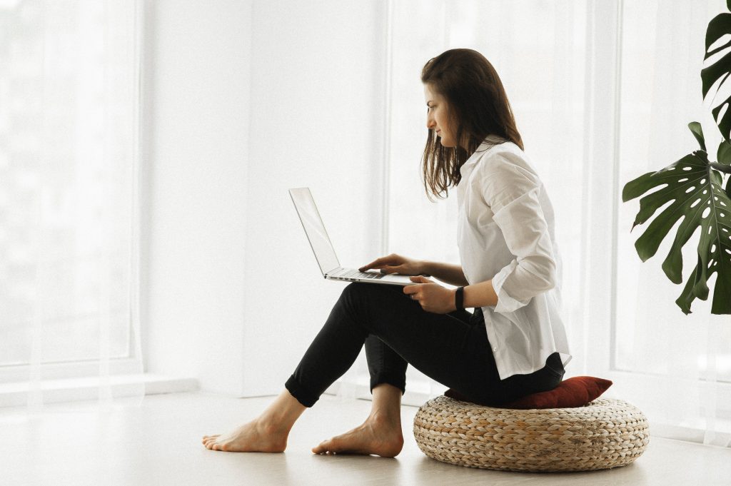 A woman sits on a woven cushion while browsing on the internet in a soft white room with lots of natural light.