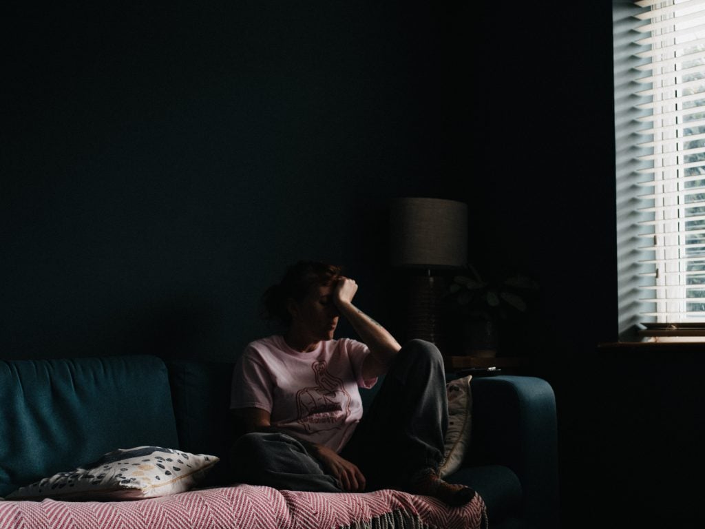 A person sitting on a couch with their head in their hand, elbow resting on their knees, looking frustrated in a dark room.