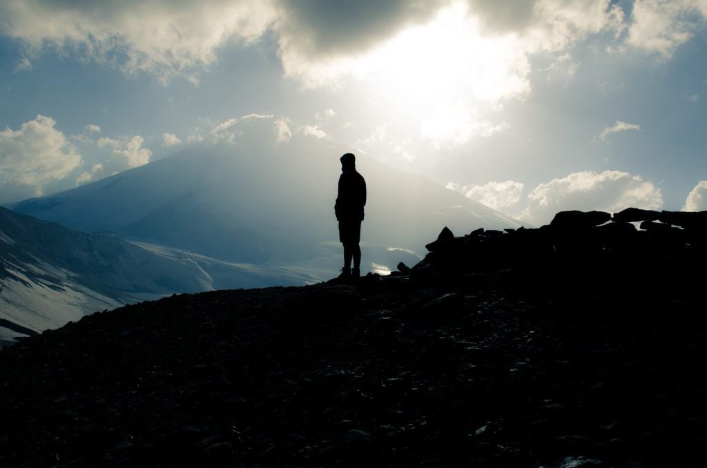 Silhouette of a man who has just climbed a mountain with the sun high in a sky of white and blue.