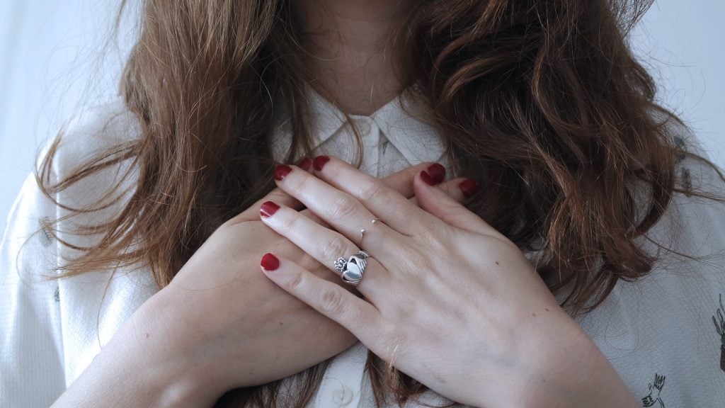 A woman holds her hands close to the collar of her shirt. She has painted red nails and is wearing a claddagh ring.