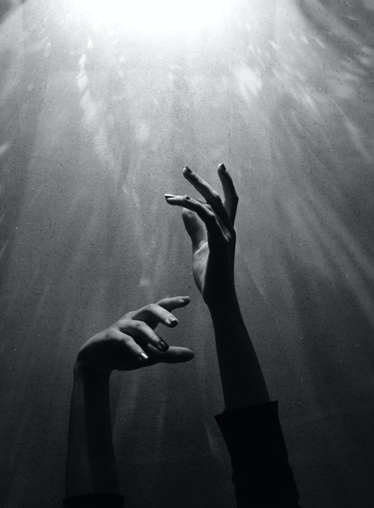A black and white photo of someone reaching up towards a light with both hands.