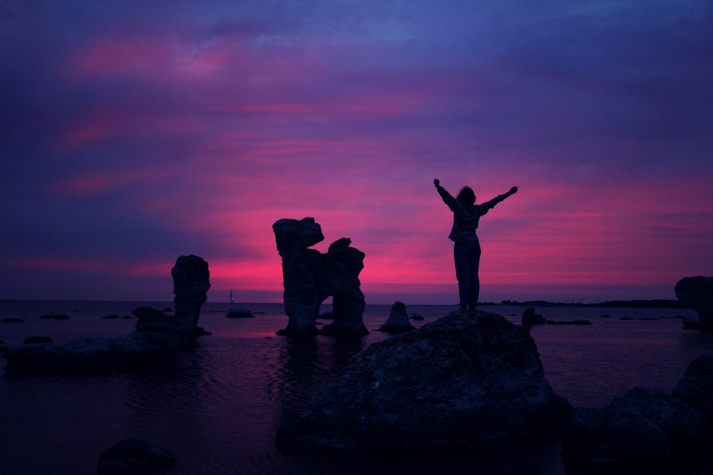 Silhouette of a person standing on a mountain with rock formations surrounding her, arms outstretched during a pink and purple sunset.