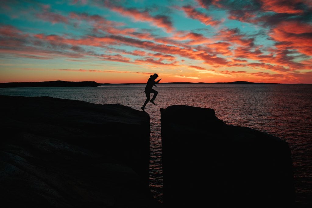 Someone jumps from cliff to cliff with a sunset over the ocean in the background.