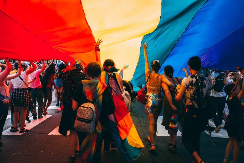 Pride parade participants march together underneath a large pride flag.