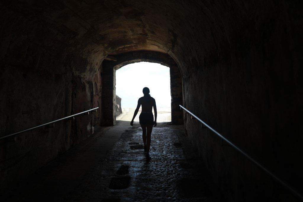 A woman walking through a dark tunnel towards the bright entrance.