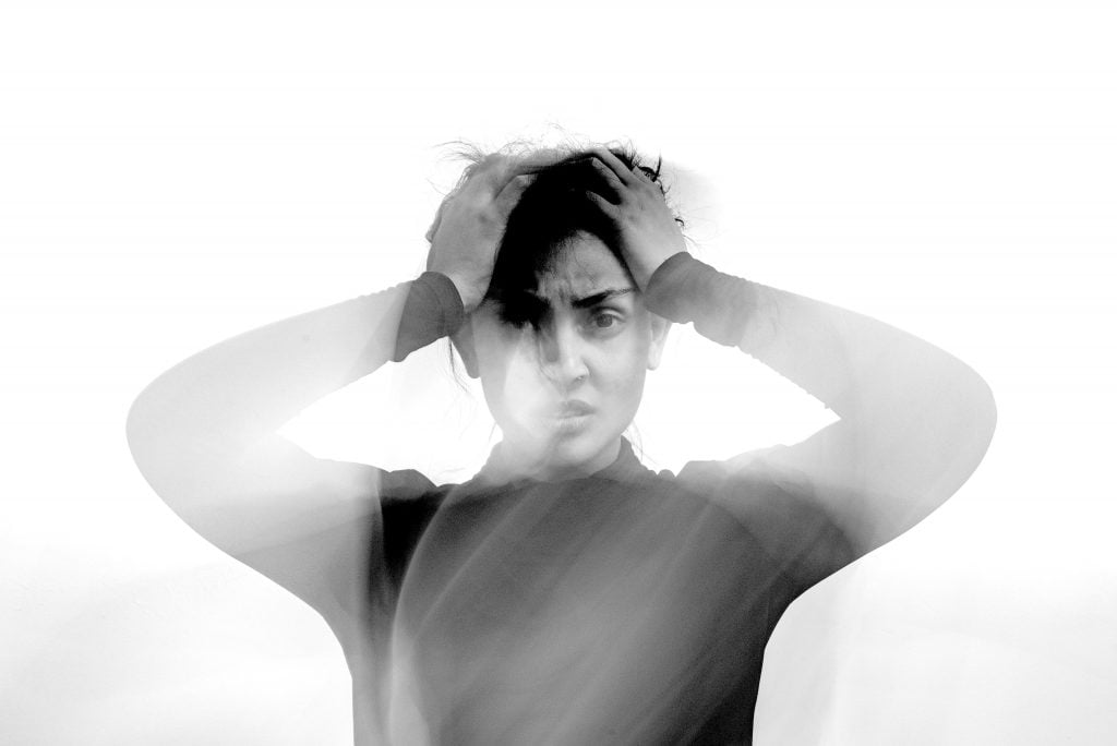 A blurry black and white photo of someone facing the camera and holding their head with both hands, looking distressed.