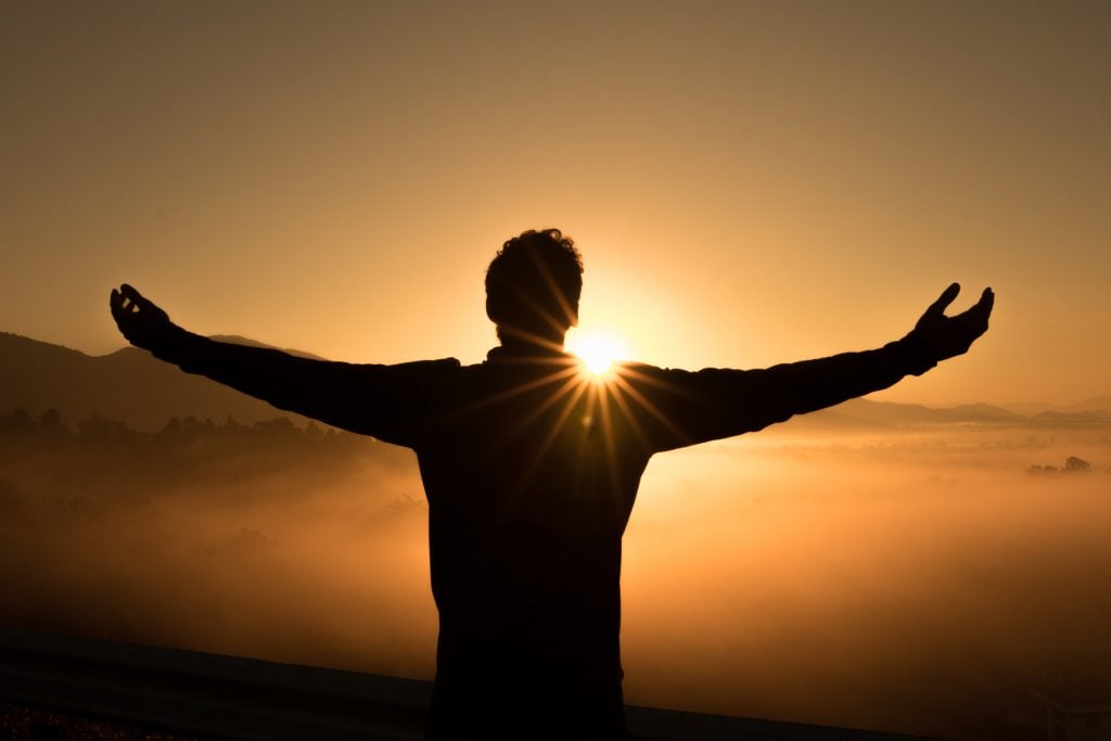 A man at the top of a mountain looks toward the golden sunrise above the clouds.