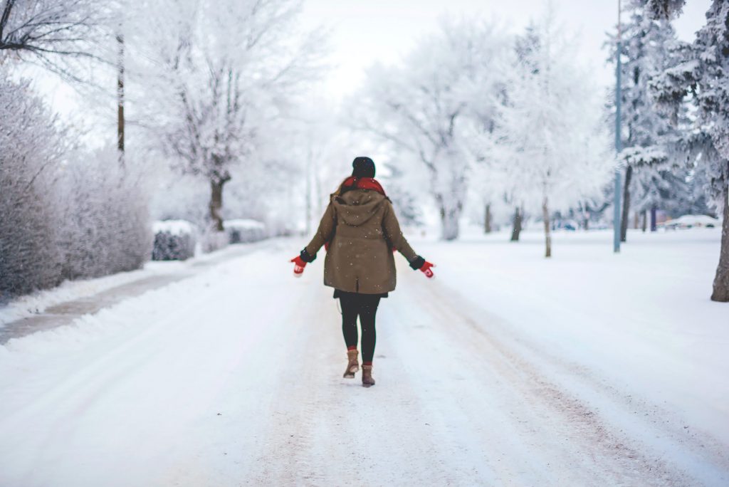 A person with their arms stretched outward walks down the road with everything blanketed in snow.