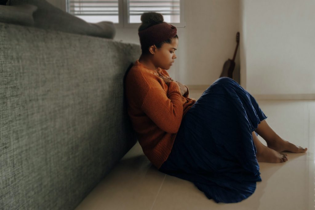 A woman sits at the foot of her bed, holding her heart, looking upset.