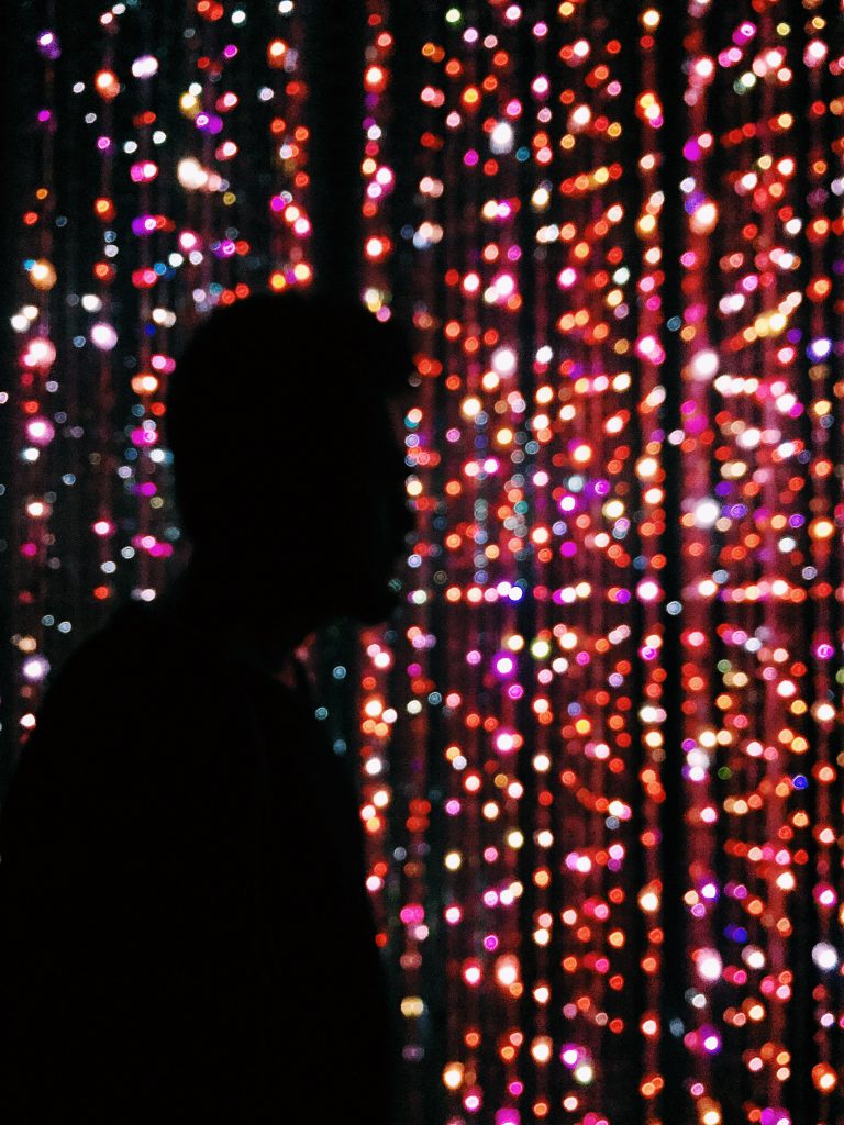 Silhouette of an individual standing in a dark room with colorful lights refracted on mirrored walls.