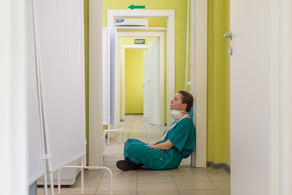 A healthcare professional sits in the hallway of a hospital. looking defeated.