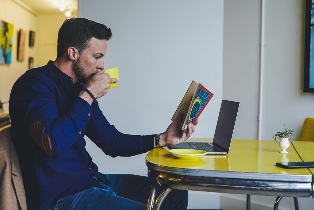 A man sits at a yellow table, drinking tea and reading a book.