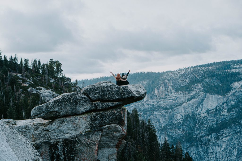 A couple sits on a rock cliff in the mountains with one arm around each other and the other arm raised with a peace sign.