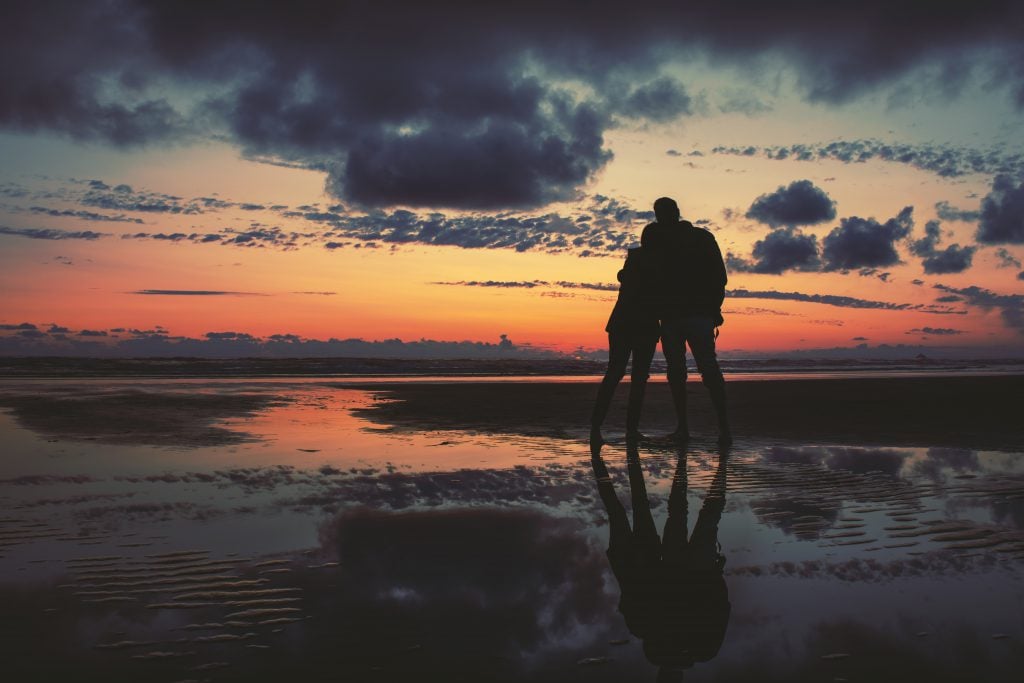 A couple stands together on a beach during sunset.