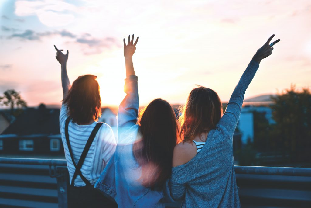 Three teenage girls facing the sunset with their arms raised in the air.