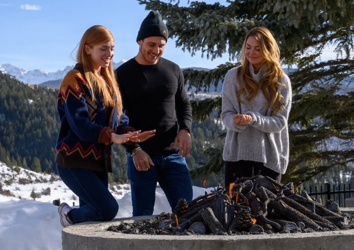 Three smiling people warm themselves by a large fire pit in the snowy Colorado mountains.