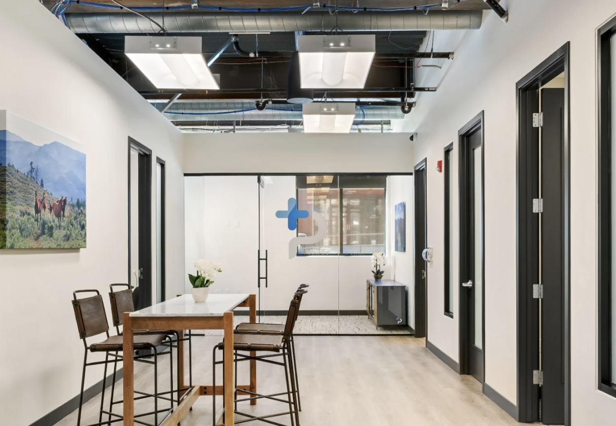 Modern office hallway with a table and chairs, featuring a landscape artwork and a glass door leading to a therapy room, reflecting the welcoming environment of the Intensive Outpatient Program in Boulder for mental health recovery.