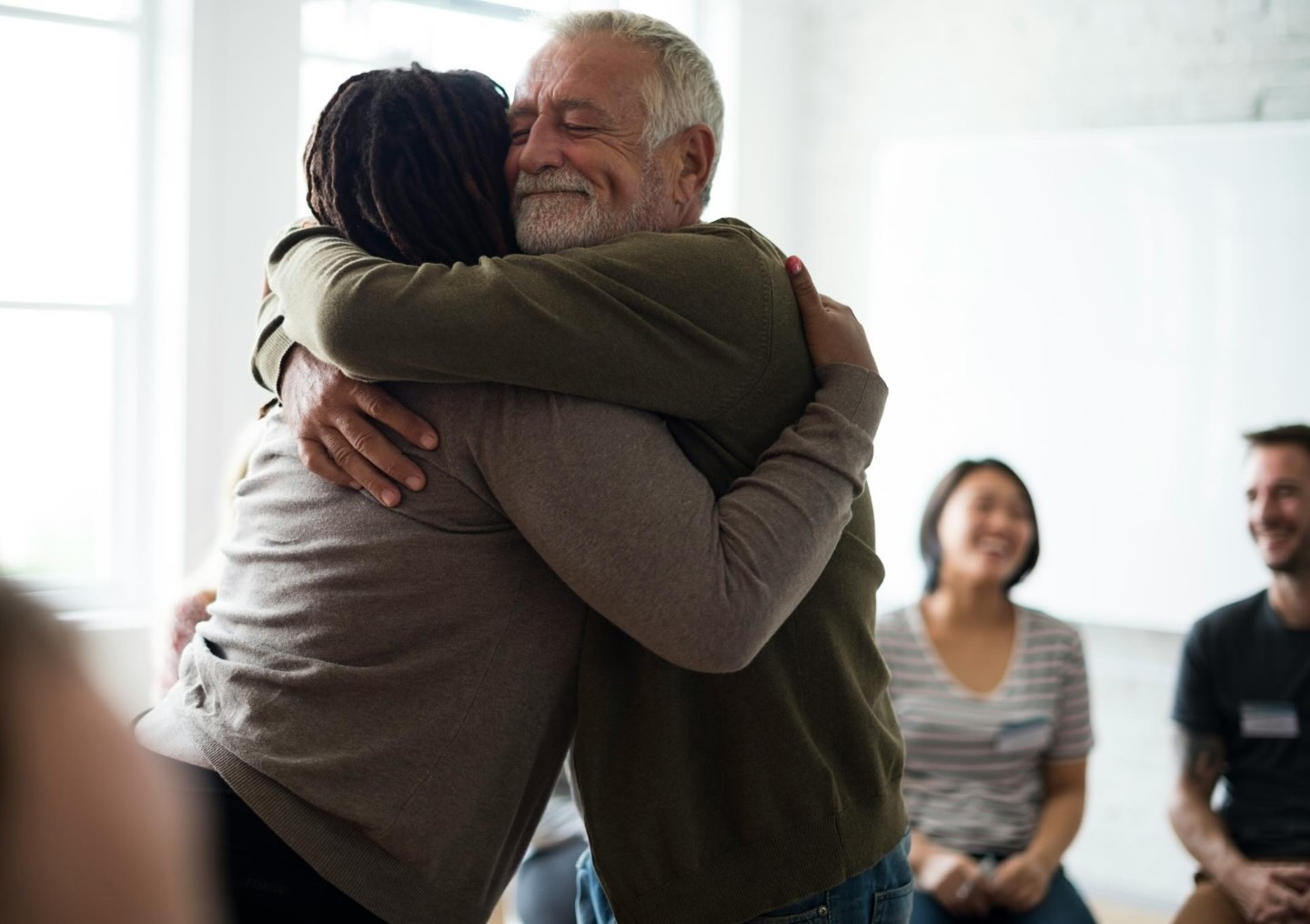 A man gently embraces another client during a group session while smiling.