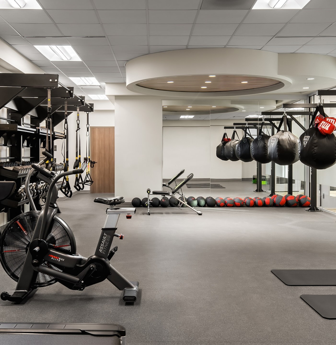9f Gym at APN Dallas-Fort Worth: an assault bike sits in front of lifting stations on the left with multiple punching bag stations on the right. A large central lighting section is framed by a circular structure on the ceiling.