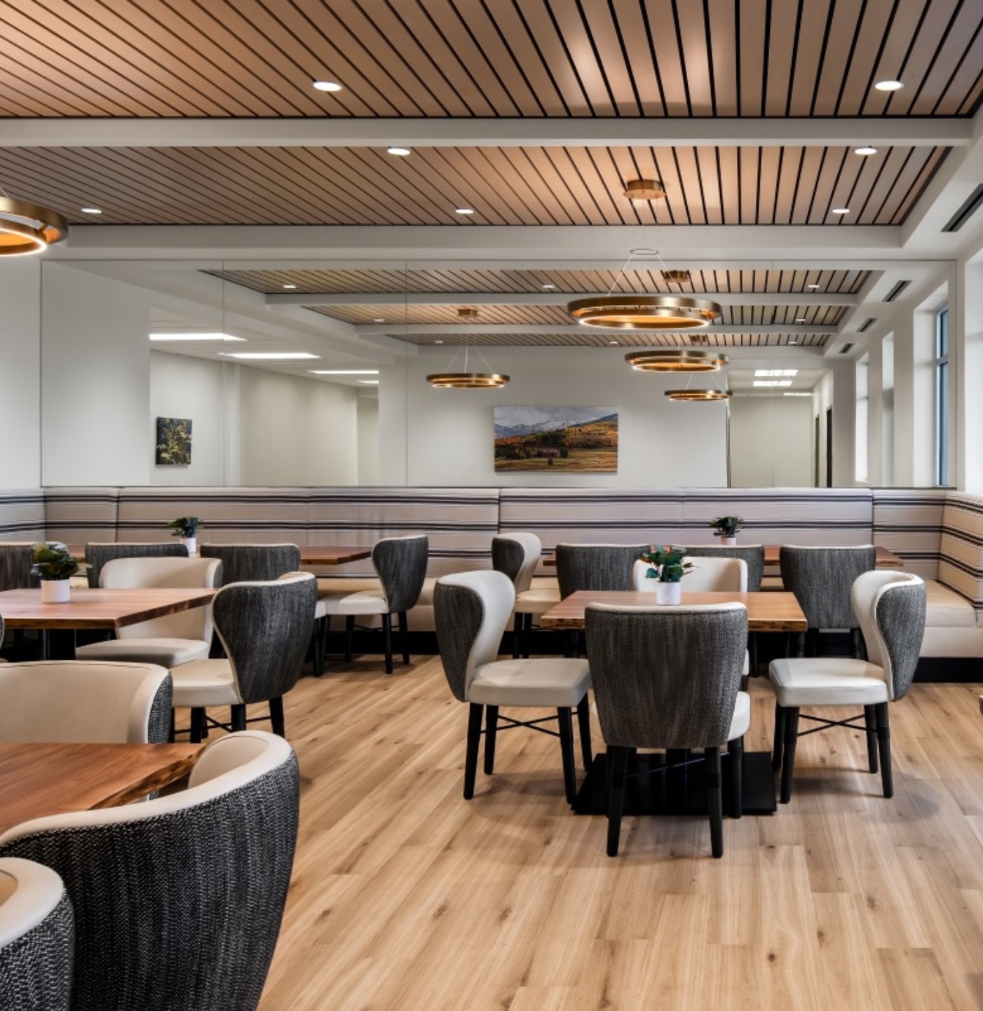 The dining room at APN Dallas-Fort Worth: natural light streams into a luxe feeling dining area with multiple table and chair groups. The ceiling is outfitted with bronze slats and recessed lighting, with a large mirror lining the back wall and reflecting lots of light.