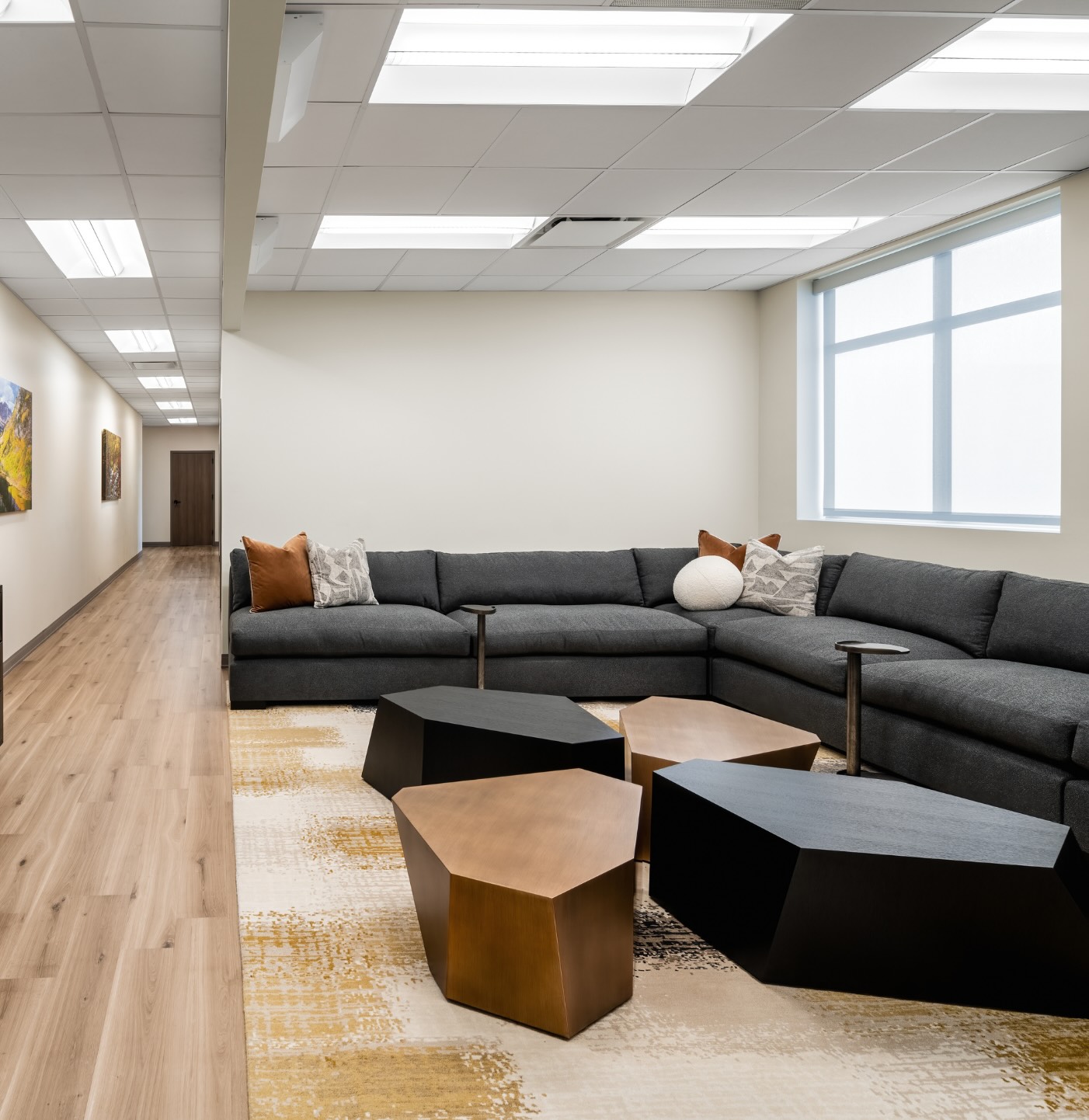The lounge at APN Dallas-Fort Worth: natural light streams into an airy seating area with large dark gray couches and geometric shaped coffee tables in dark and light wood grains.