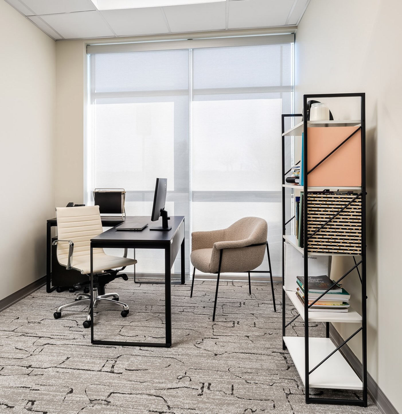 A therapy room at APN Dallas-Fort Worth: the room is backlit by a large set of windows filling the whole wall with a sheer white blind. Two chairs sit on either side of a desk with a bookcase lined with different office supplies and decor.