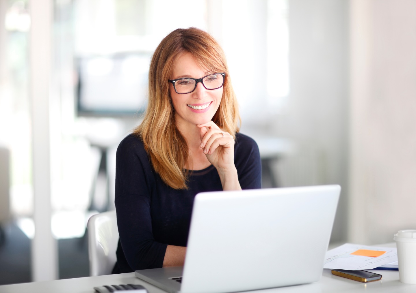 A woman wearing glasses sits at a desk with a white laptop. She has her hand on her chin and is smiling while requesting information online.