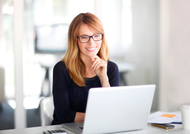 A woman wearing glasses sits at a desk with a white laptop. She has her hand on her chin and is smiling while requesting information online.