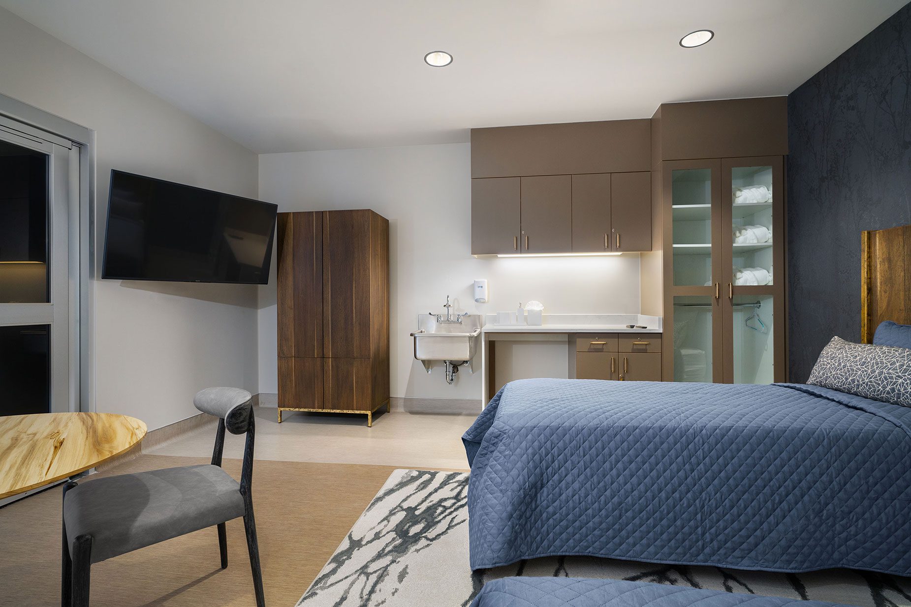 Luxury treatment room at All Points North featuring a blue quilted bedspread, modern furnishings, a television, and upscale amenities for holistic recovery.