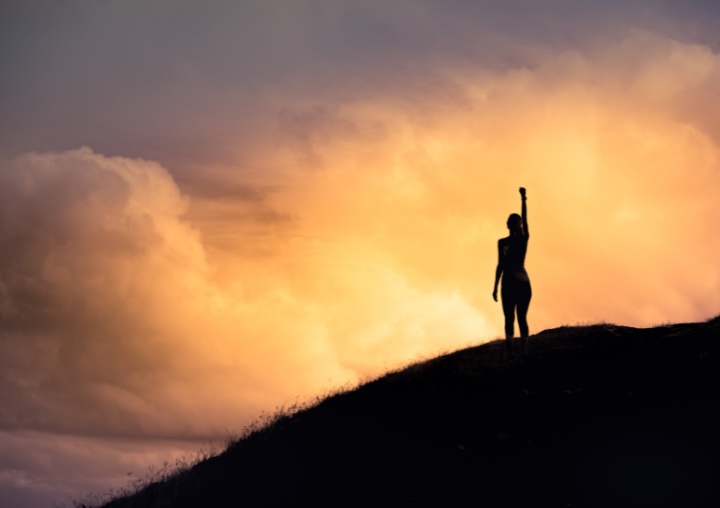 A person stands on the top of a hill at sunset with their fist in the air.