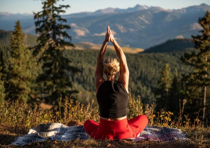 A woman sits in a yoga pose with her arms extended over her head and her hands pointing up with a mountain scene in front of her.